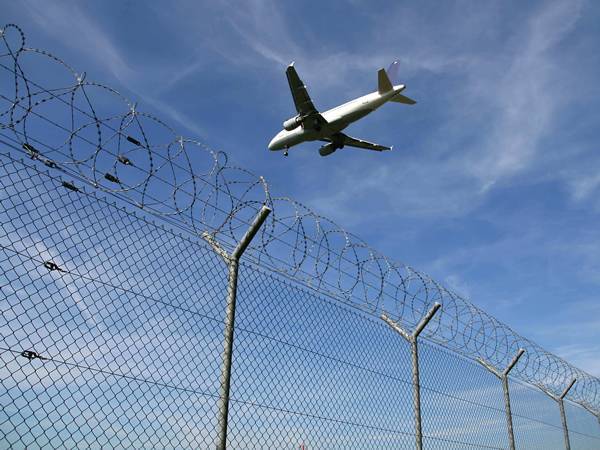 An airplane is flying over the chain link fence equipped with razor wires.