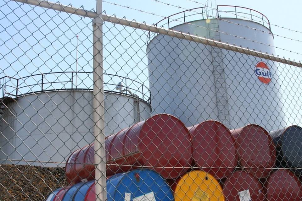 Several oil tanks are stored in the yard enclosed by chain link fence with barbed wire.